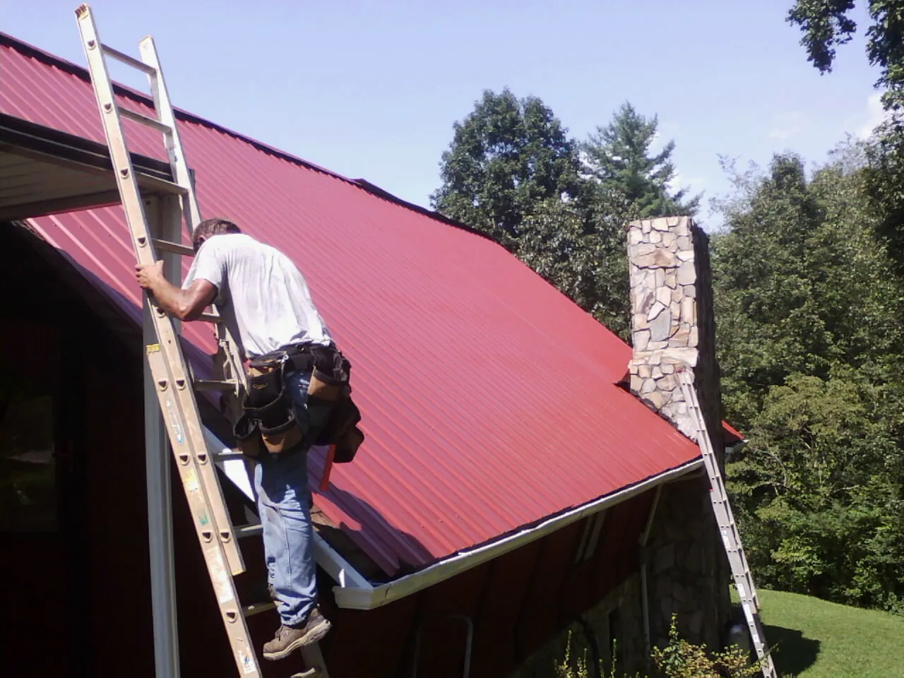 Roofer on a ladder beside a red metal roof and stone chimney — Soffit & Fascia Repair by Roofing Brothers in Bell Gardens